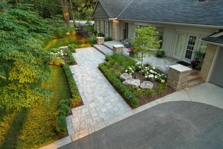 A front walkway made of stone that is bordered by lush gardens and square hedges.