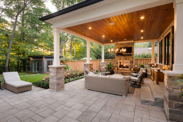 Cozy outdoor patio with seating, a fireplace, and wooden ceiling. Surrounded by trees, flowers, and a well-maintained lawn, creating a serene backyard setting.