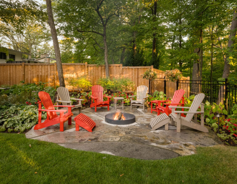 A cozy backyard scene features vibrant Adirondack chairs surrounding a fire pit, enclosed by a wooden fence, and lush greenery in the background.