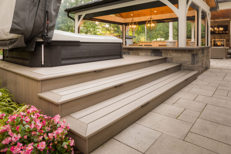 A cozy outdoor patio with a covered seating area, hot tub, wooden steps, and vibrant flowers. Warm lighting creates an inviting atmosphere.