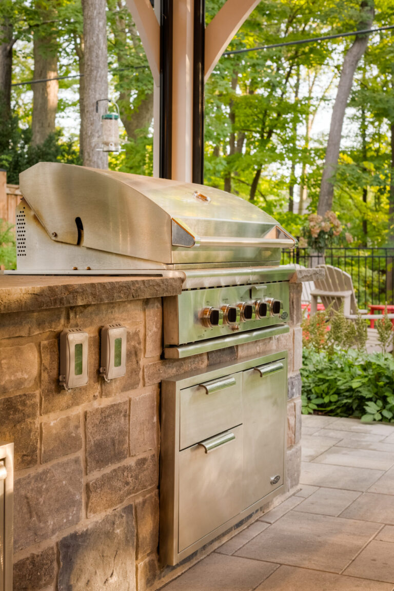 A modern outdoor kitchen features a stainless steel grill on a stone counter, surrounded by lush greenery and a cozy patio setting.