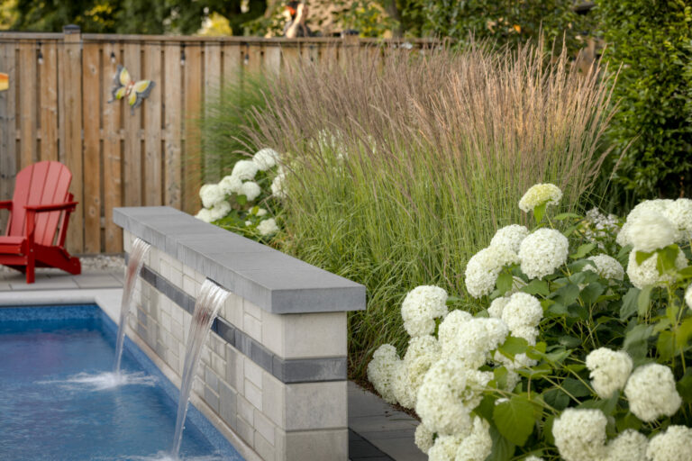 A backyard pool with cascading water feature, red chairs, tall grasses, white hydrangeas, and a wooden fence adorned with a butterfly decoration.