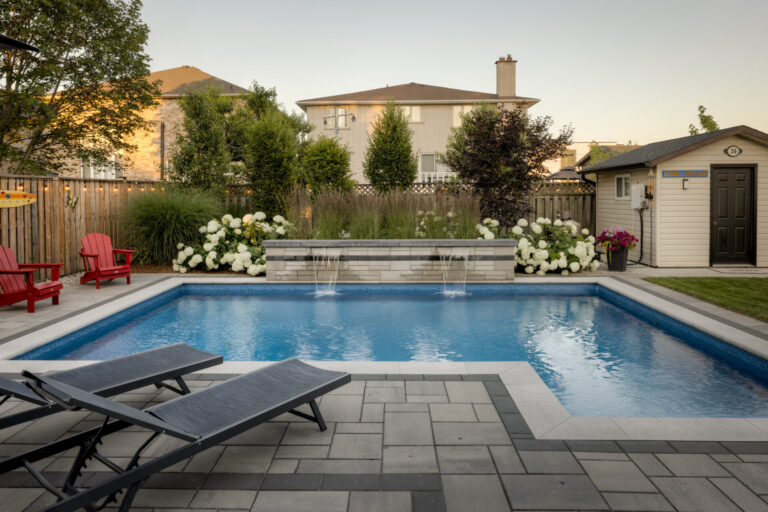 Backyard with a swimming pool, red chairs, and lounge chairs. Surrounded by plants and a wooden fence. Small shed visible in the background.