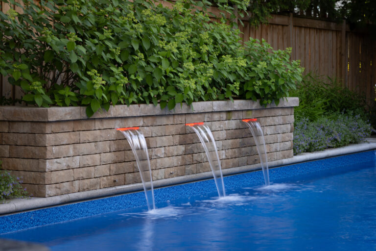 A backyard pool with three water spouts flows elegantly. Surrounded by lush greenery and a wooden fence, offering a private, serene atmosphere.