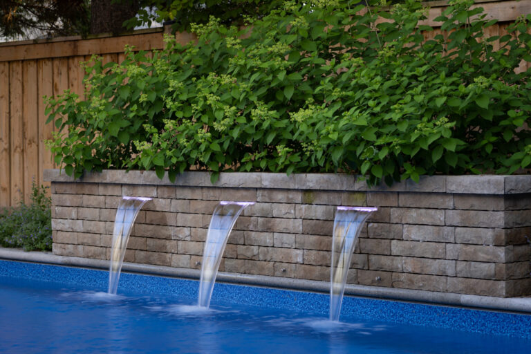 Three elegant water fountains flow into a serene blue pool, bordered by a lush green hedge and a wooden fence, creating a relaxing ambiance.