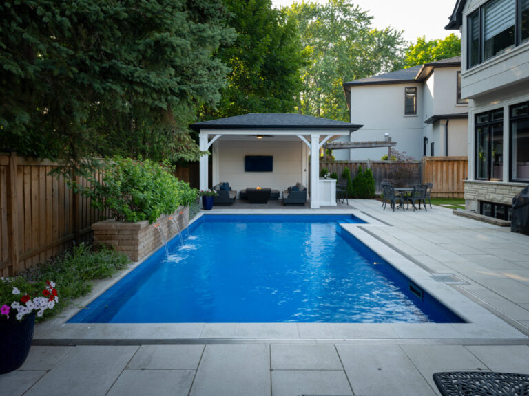 A backyard features a rectangular swimming pool, patio area, seating under a gazebo, and tall trees along a wooden fence. No people present.