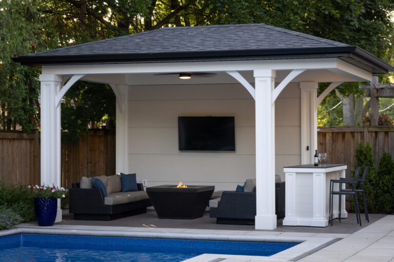 A poolside pavilion with a flat-screen TV, seating area, and bar sits near a blue-tiled pool, surrounded by trees and a fence.