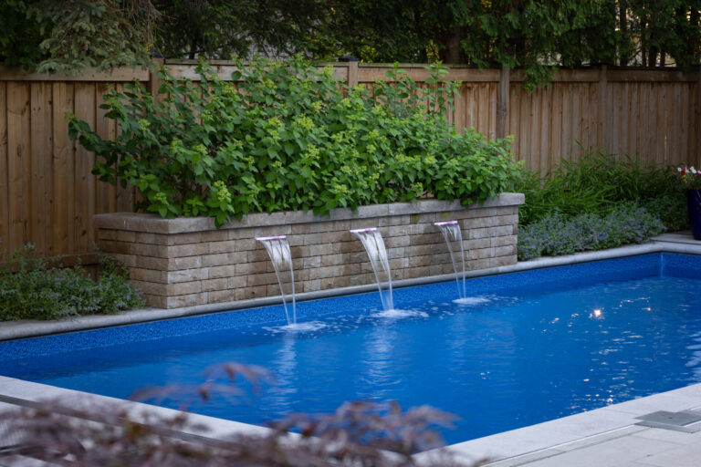 A backyard swimming pool with three small waterfalls from a brick wall, surrounded by green plants, beside a wooden fence.