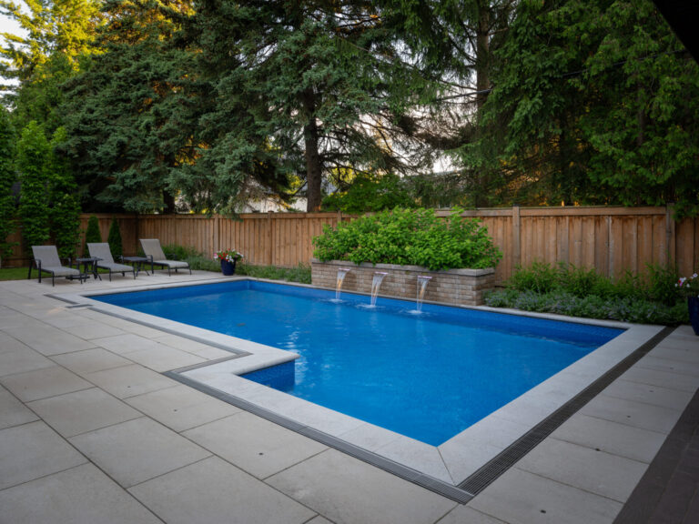 A modern rectangular swimming pool with water features, surrounded by a wooden fence, lounge chairs, and tall evergreen trees in a serene backyard setting.