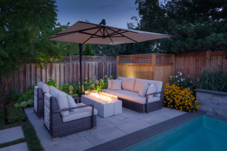 Outdoor patio scene with wicker furniture, a large umbrella, and a lit central firepit beside a pool, surrounded by plants and wooden fencing.