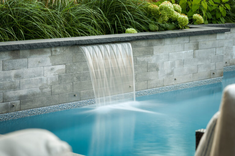 A serene pool with a waterfall feature flows over a stone wall, surrounded by lush green plants, creating a tranquil outdoor setting.