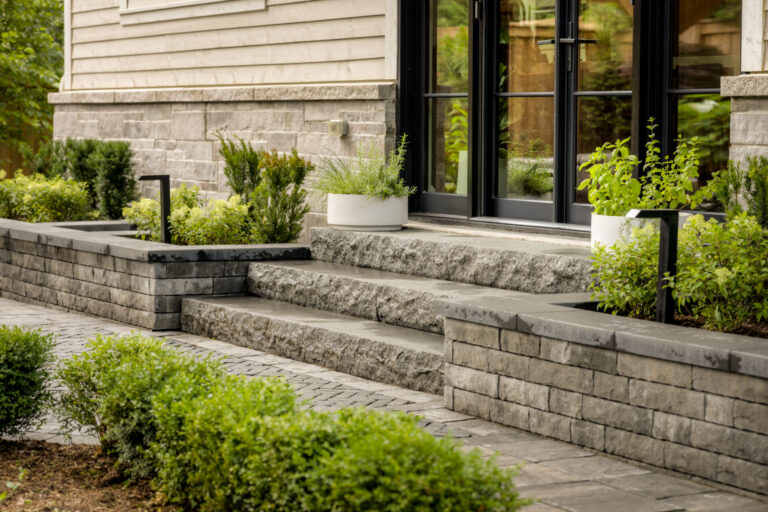 Modern home entrance with stone steps, planters, and large glass doors. Trimmed bushes line the path, creating a clean and inviting entrance.