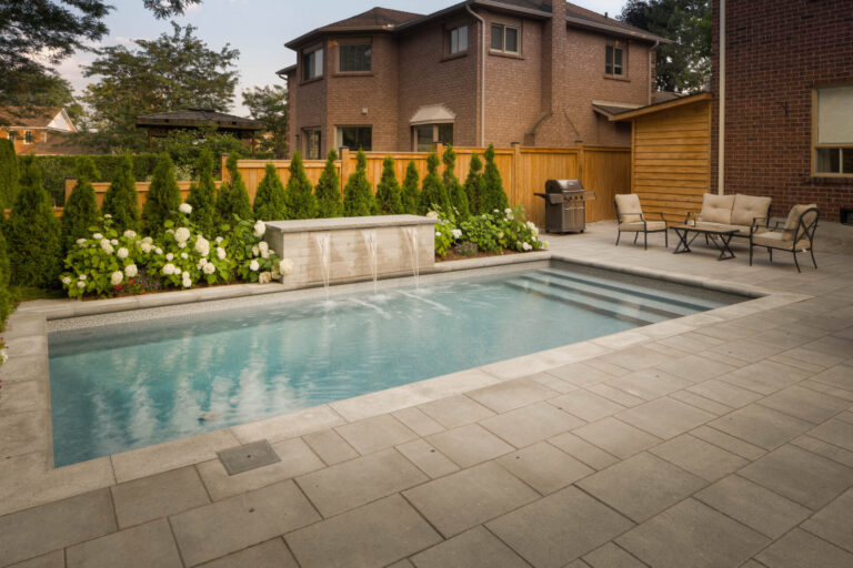 A backyard features a small pool, patio furniture, and a barbecue grill, surrounded by greenery and fenced by wooden panels.