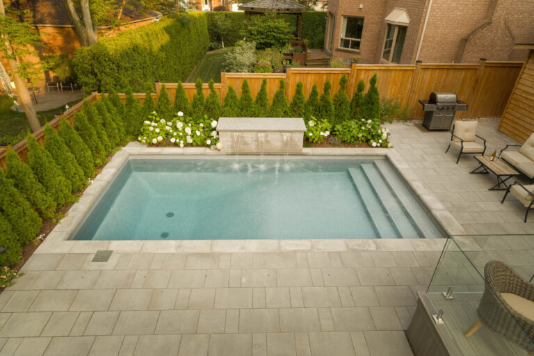 A small backyard pool with a fountain, surrounded by patio furniture and greenery, near a brick house and wooden fence. No people visible.