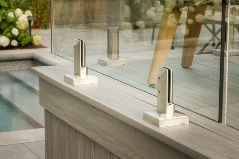 Glass railing with metal brackets surrounds a pool area, featuring wooden decking and flowers in the background. Peaceful, modern outdoor setting.