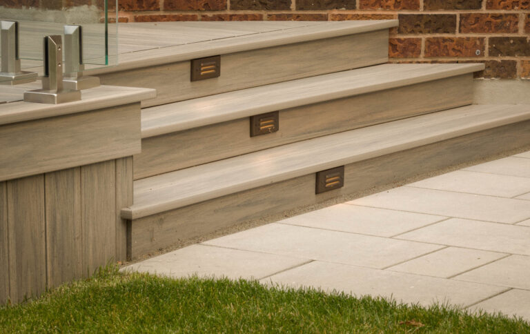 Modern wooden steps with built-in lighting lead to a brick wall. Adjacent are glass railings and a neatly maintained lawn.