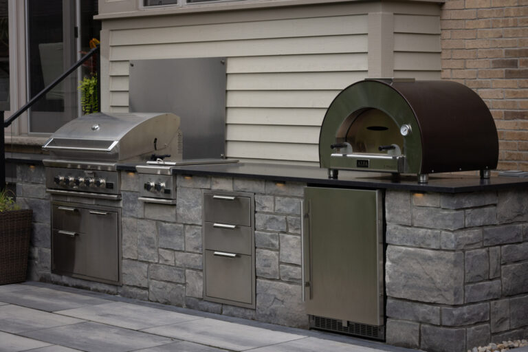 Outdoor kitchen with stainless steel grill, pizza oven, and storage, set against a stone countertop. Modern residential patio setting in background.