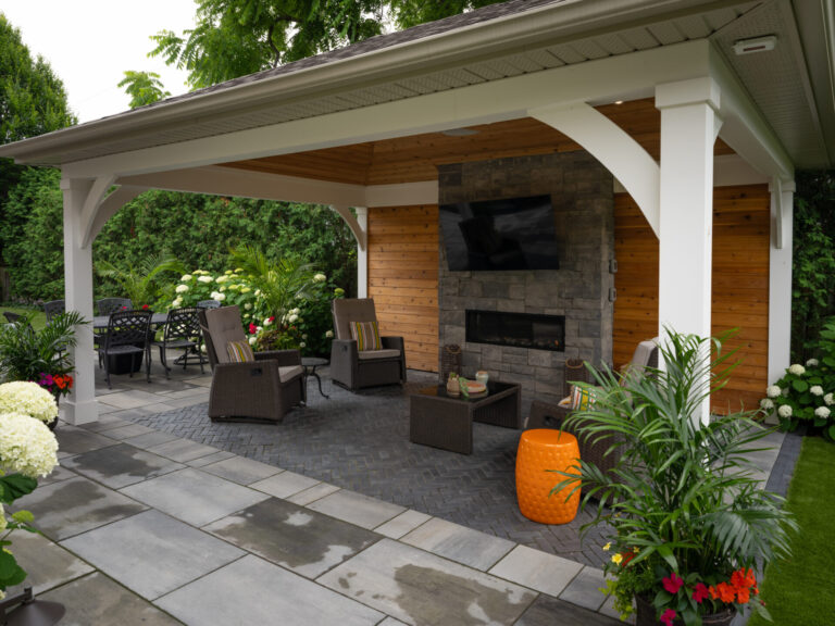 A modern outdoor patio features a gazebo with a TV, fireplace, wicker chairs, lush greenery, and vibrant flowers, creating a cozy relaxation area.