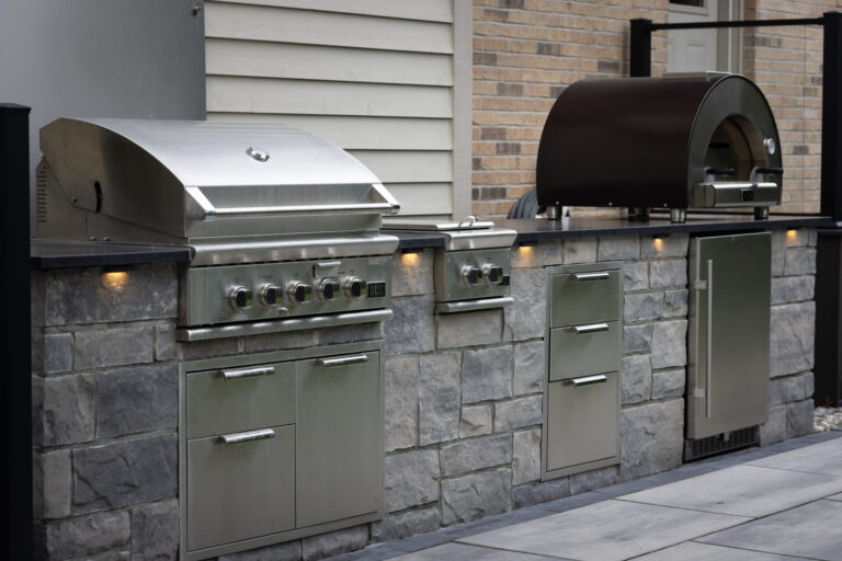 The image shows a modern outdoor kitchen with stainless steel appliances, including a grill and oven, set against a stone and brick wall.