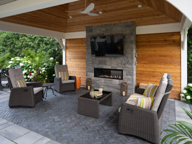 Covered patio with wicker furniture, stone fireplace, and television. Surrounded by greenery and flowers, creating a cozy outdoor living space with a modern feel.