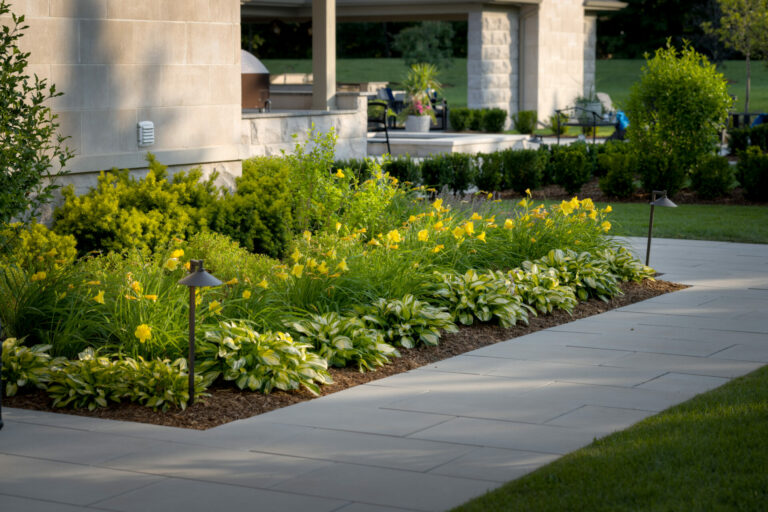 A modern garden features neatly arranged yellow flowers and lush greenery alongside a paved walkway, leading to a patio with outdoor seating.