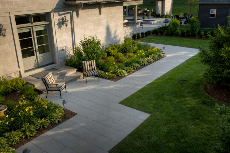 A well-maintained garden patio with chairs, lush greenery, and paved walkway adjacent to a building, extending to a grassy area with shrubs.