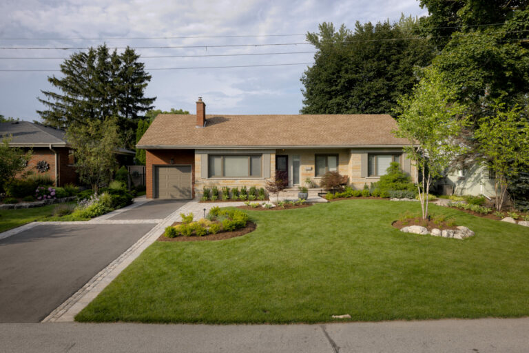 Suburban house with a well-kept lawn, surrounded by trees and shrubs. A driveway leads to the attached garage. Clear skies above.