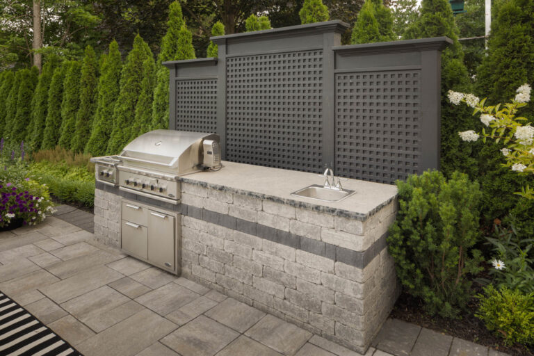 Outdoor kitchen with a granite countertop featuring a grill and sink. Surrounded by lush greenery and modern lattice fencing. Paved patio floor.