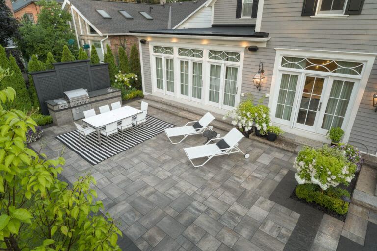 Spacious backyard with a modern patio setup, white dining table, chairs, striped rug, lounge chairs, and surrounding greenery in a residential setting.