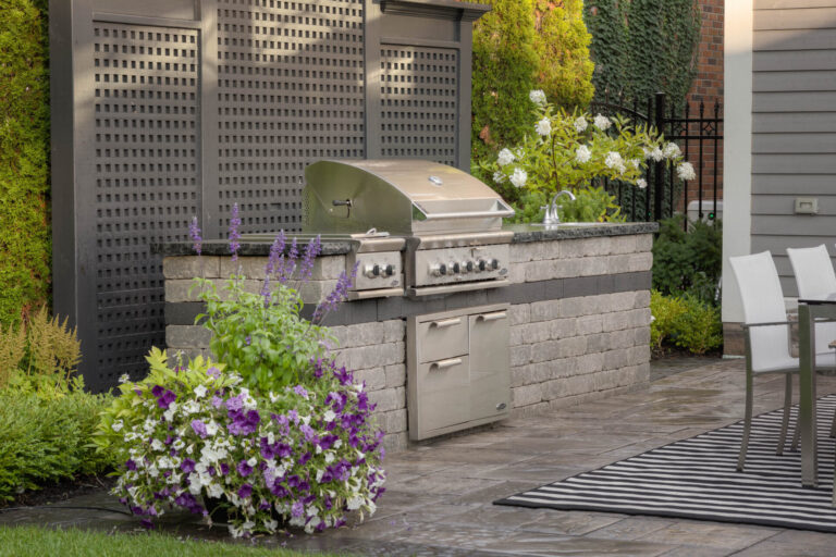 Outdoor patio featuring a stainless steel grill, surrounded by lush greenery and colorful flowers, adjacent to a modern seating area with a rug.