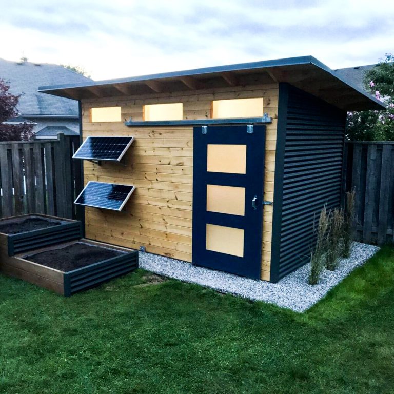 A standalone, outdoor room with solar panels, a barn door, wood siding, and two empty flower beds.