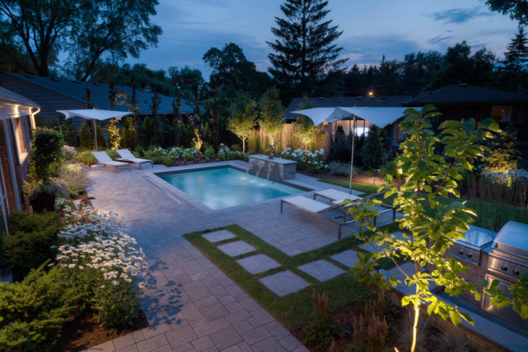 A beautiful inground pool with water features is surrounded with patio stones, beach chairs, and umbrellas. It is night time.