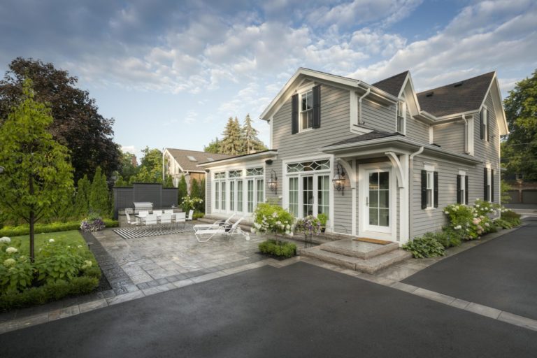 A house with a large driveway and adjoining front stone patio with a grill, dining area, and sun bathing chairs.