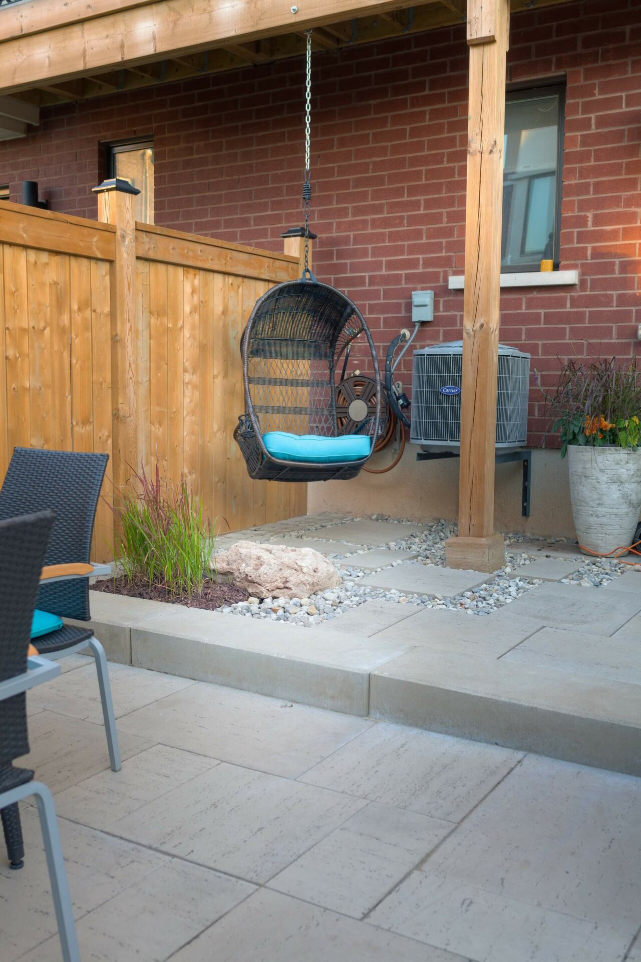 A cozy backyard patio with a hanging chair, wooden fence, potted plants, and brick wall; ideal for relaxing and outdoor gatherings.