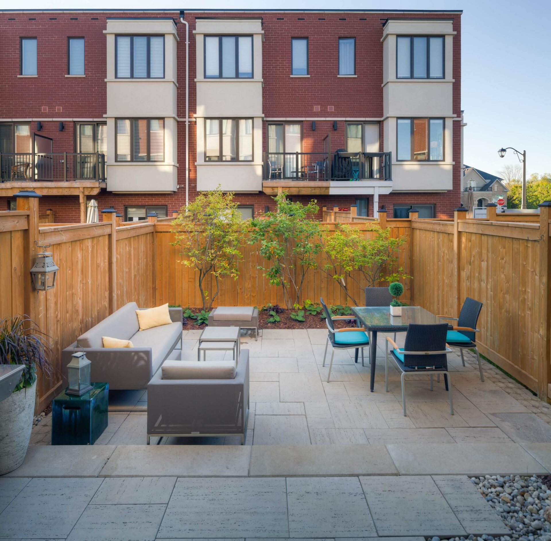 Enclosed backyard patio with modern outdoor furniture, bordered by wooden fencing. The view includes brick townhouses in the background, and potted plants.