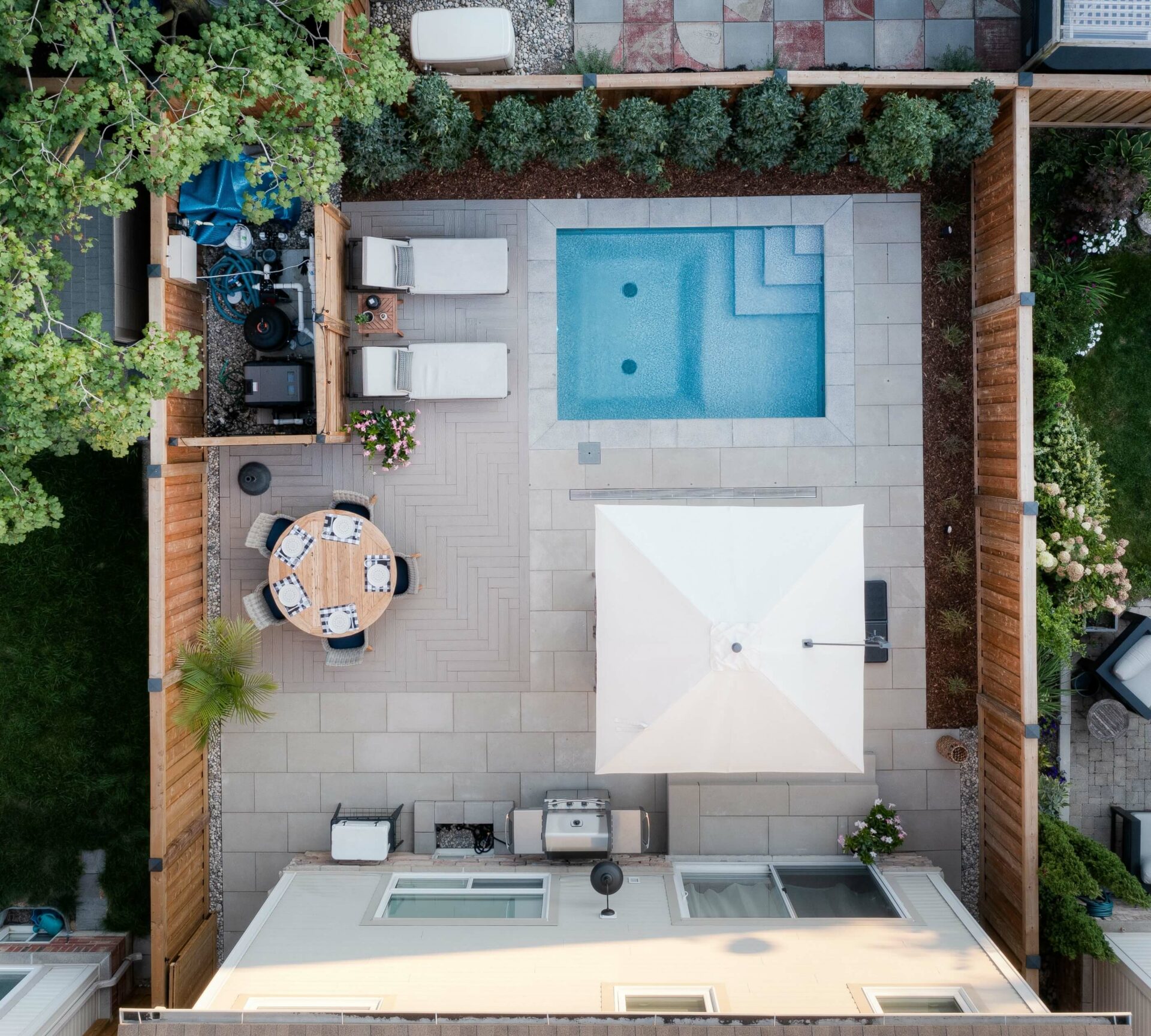 Aerial view of a backyard with a rectangular pool, patio area, round table, umbrella, barbecue, and surrounding plants.