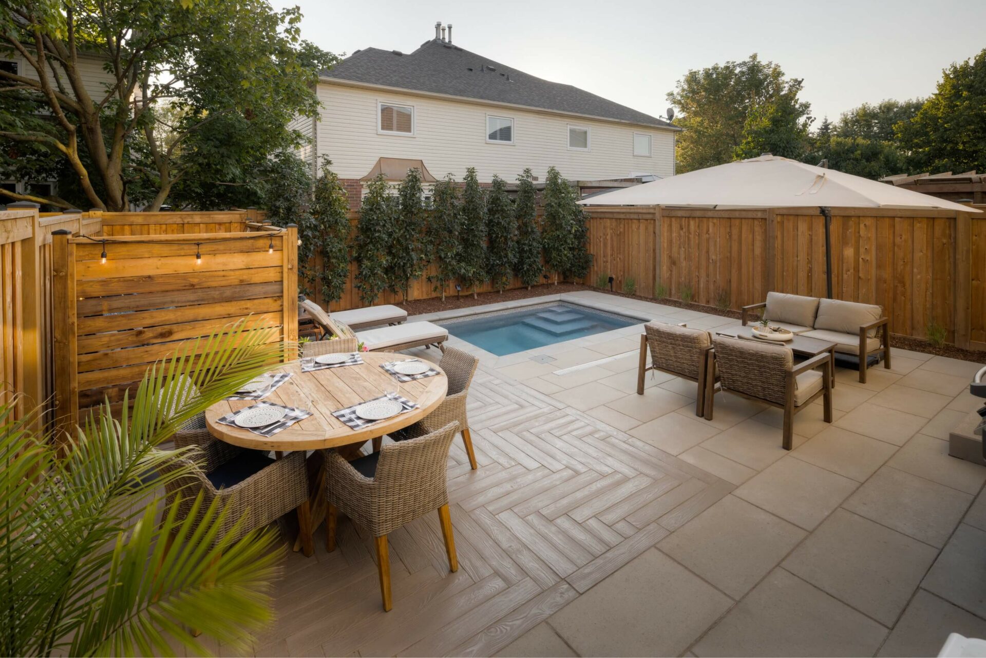 A cozy backyard features a small pool, patio furniture, and wooden fencing, with two-story houses and lush greenery surrounding the area.