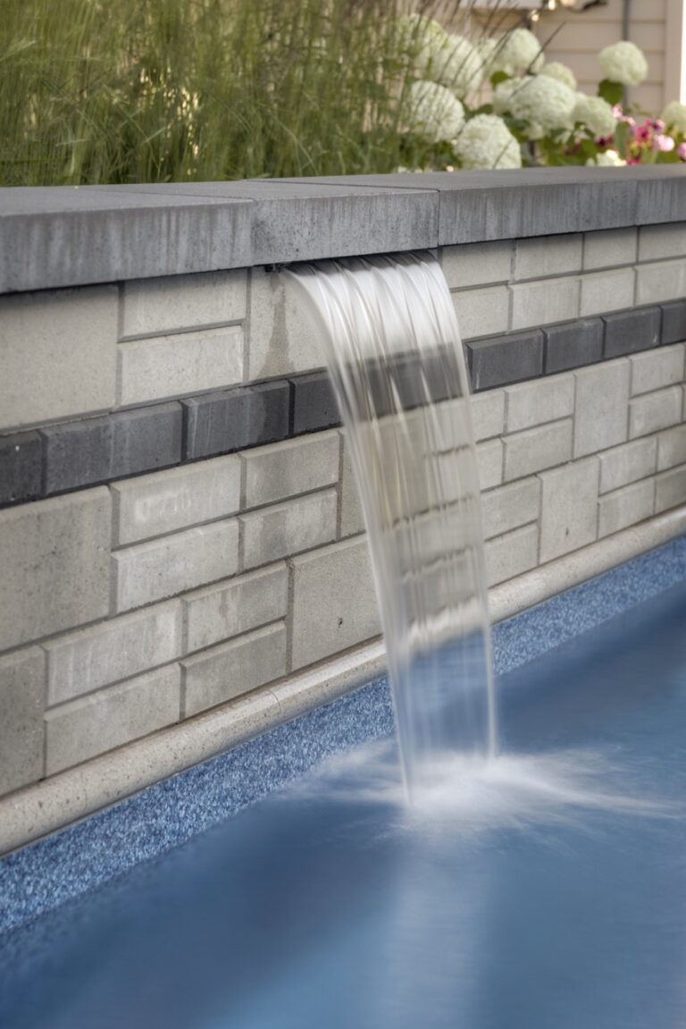 Water cascades from a stone wall into a pool, surrounded by greenery and flowers, creating a tranquil, modern garden scene.