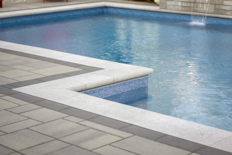 A serene swimming pool corner with clear blue water and light-gray stone tiles, creating a peaceful and inviting atmosphere. No people are present.