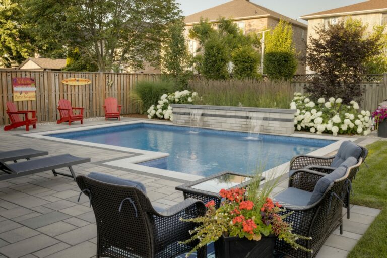 A backyard with a rectangular pool, surrounded by wicker chairs, colorful flowers, and lush greenery, creates a tranquil outdoor retreat.