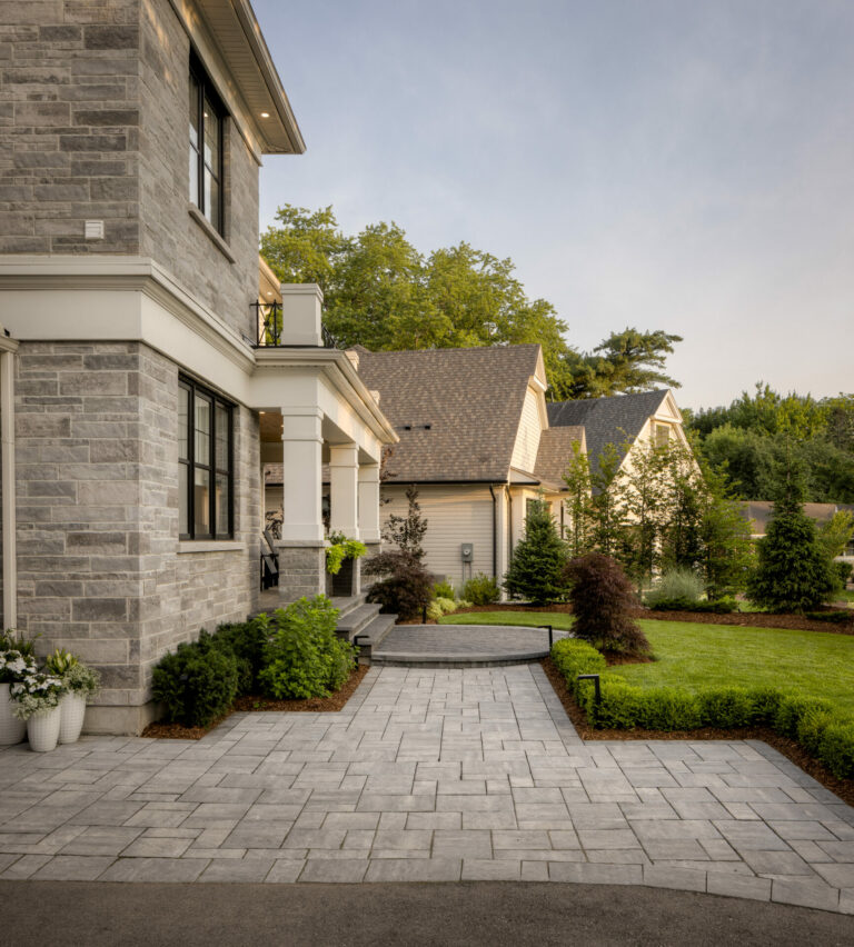 Elegant stone house with a well-maintained garden, potted plants, and a paved driveway. Bright, sunny day enhances the serene residential setting.