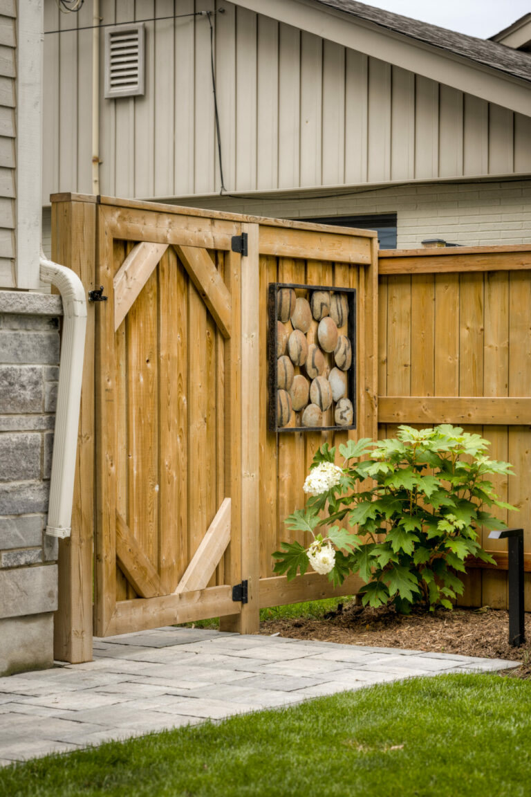 A wooden gate with decorative stones stands beside a house. A small flowering plant grows nearby on a neatly manicured lawn.