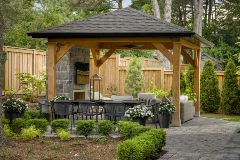 A wooden gazebo in a landscaped backyard, featuring a stone fireplace and outdoor seating surrounded by lush greenery and a wooden fence.