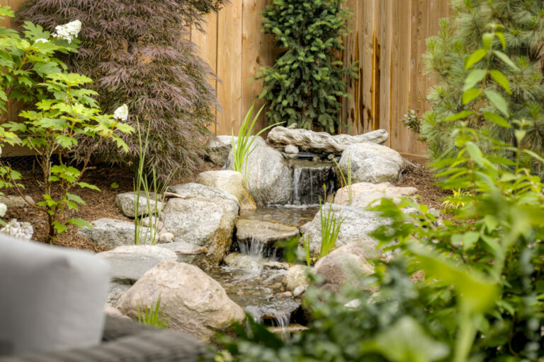 A tranquil garden scene featuring a small rock waterfall surrounded by lush greenery and a wooden fence, creating a serene, natural ambiance.