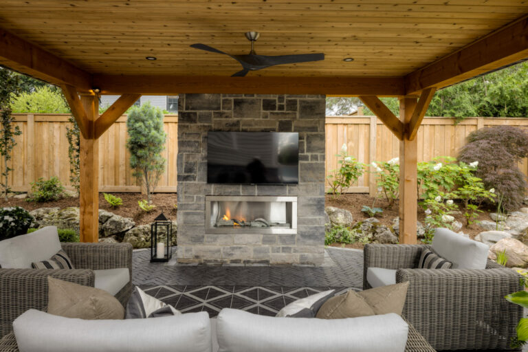 Outdoor patio with cozy seating, stone fireplace, and TV. Covered by a wooden pergola. Lush greenery surrounds the space, creating a private oasis.