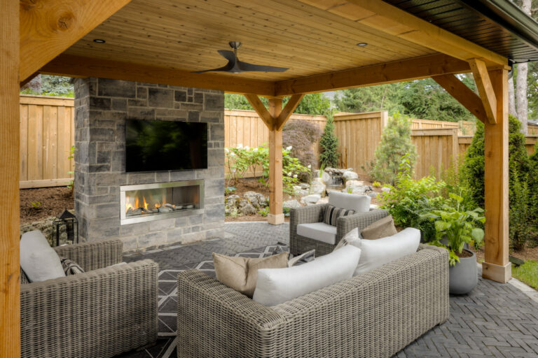 A cozy outdoor patio features wicker seating, a stone fireplace, and a TV under a wooden pergola. Lush greenery surrounds the fenced area.