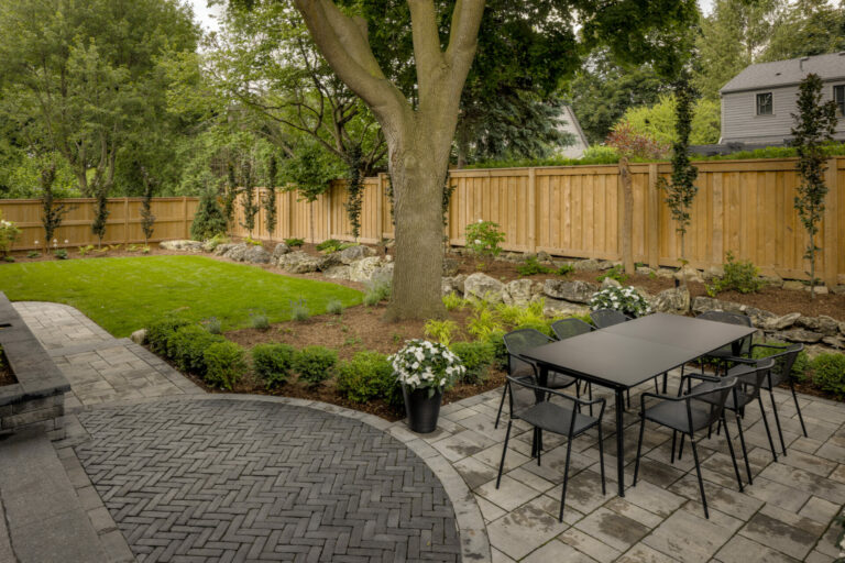 A serene backyard features a patio with a dining table, surrounded by lush greenery, a tree, and a wooden fence.