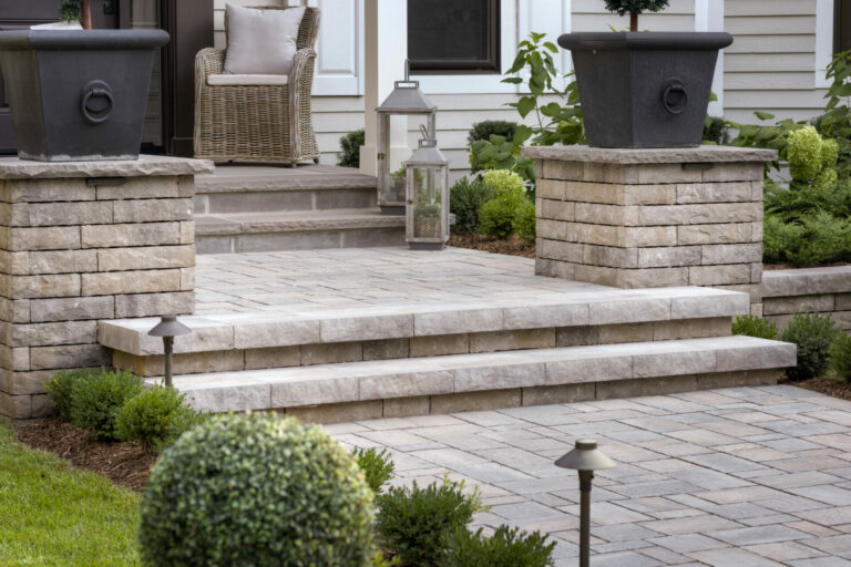 Stone steps lead to a porch with wicker chair, large planters, and lantern. Neatly trimmed bushes and pathway lights frame the entrance.