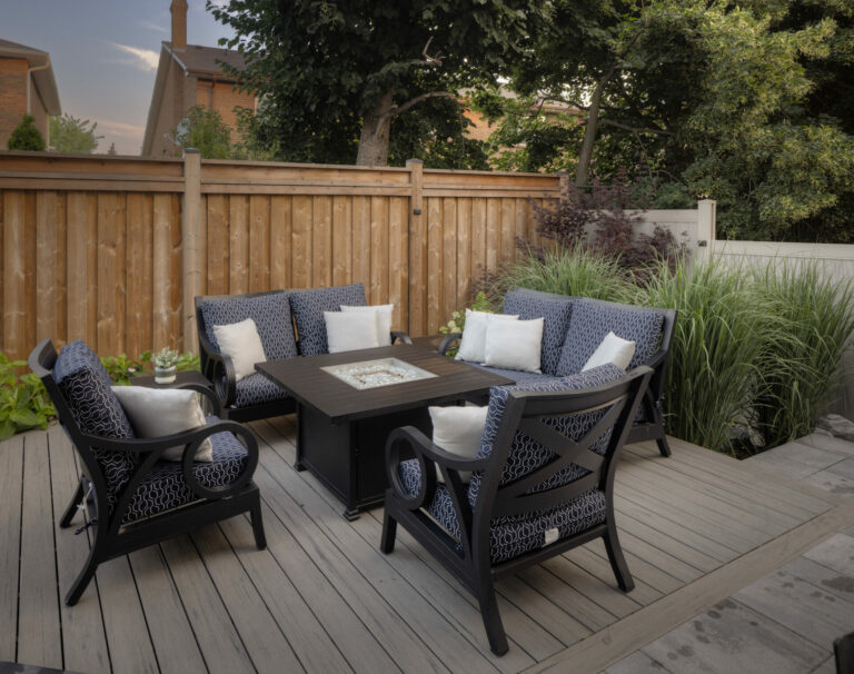 Outdoor seating area with four chairs around a table on a wooden deck, surrounded by plants and a wooden fence.