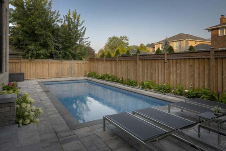 A backyard features a rectangular swimming pool, surrounded by a wooden fence, with patio chairs and lush greenery under a clear sky.